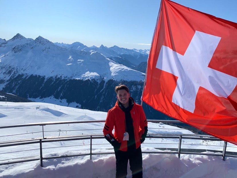 Dobin-Smith smiles next to flag of Switzerland during a quick exploration of the Swiss Alps. The group house he would share with other participants of the Wisdom Accelerator for Youth program is located across the valley.