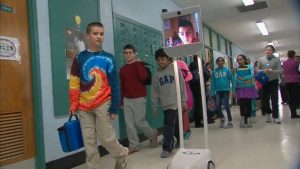Students walk in hallways to get to class as usual, while their peer who was out sick is wheeled around on a rolling screen. This student was able to keep up in class and is another futuristic alternative to keep kids up to date when they have to miss class.
